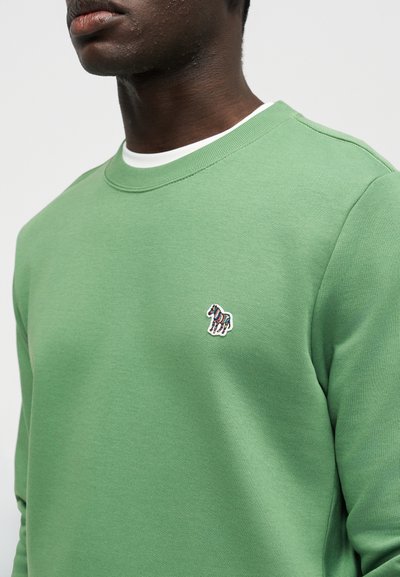 Άνδρας που φοράει ανοιχτοπράσινο φούτερ πάνω από λευκό πουκάμισο, με ένα μικρό πολύχρωμο λογότυπο ζέβρας στο στήθος.