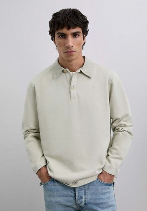 Camisa polo de manga larga gris claro con un cuello clásico, que presenta una tapeta de tres botones y un sutil detalle de logo. Combinada con unos vaqueros.