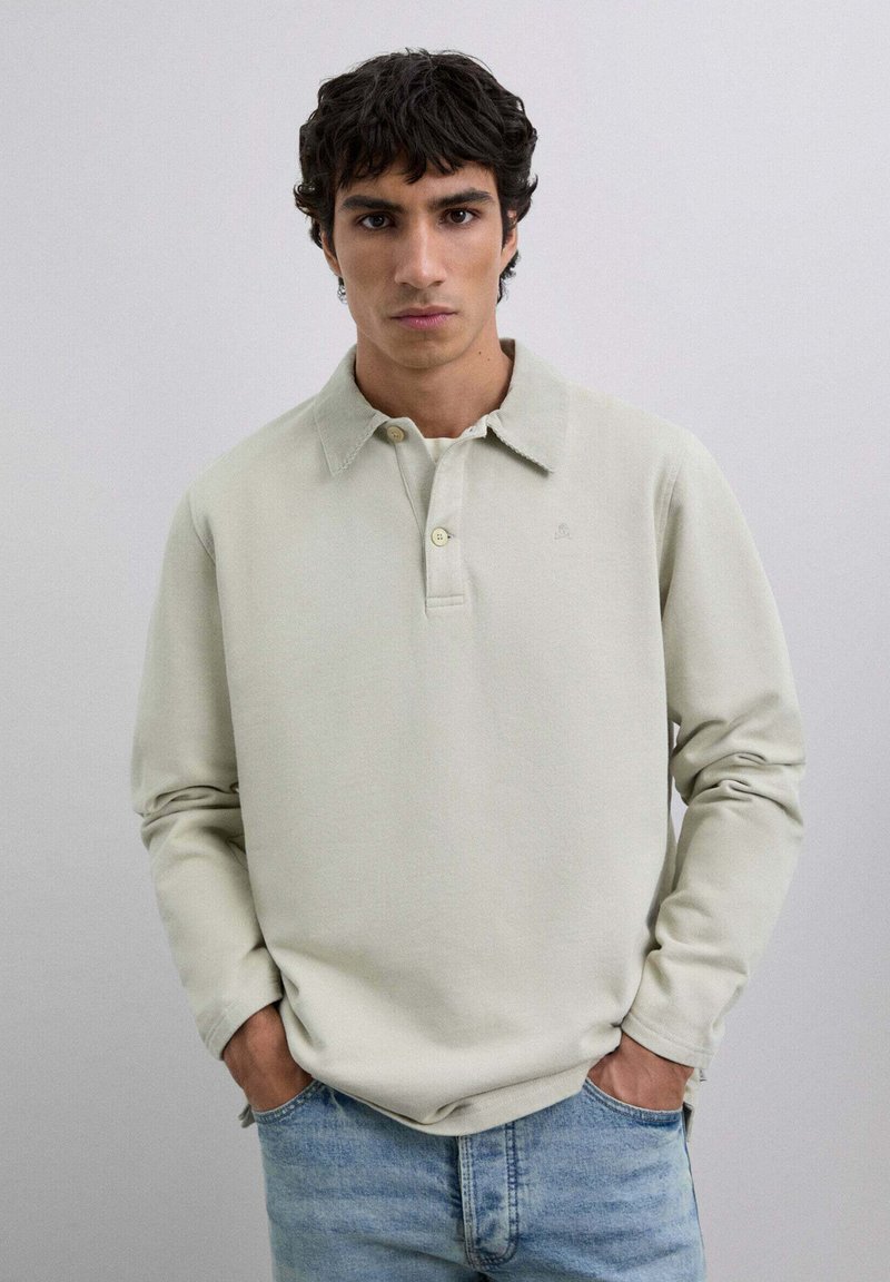 Camisa polo de manga larga gris claro con un cuello clásico, que presenta una tapeta de tres botones y un sutil detalle de logo. Combinada con unos vaqueros.