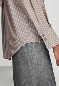 Personne portant une chemise à manches longues rayée taupe avec des poignets boutonnés et un pantalon gris texturé, debout devant un fond uni.