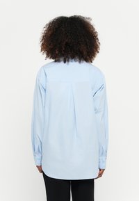 Camisa de manga longa azul claro, de corte solto, com uma preguiça nas costas, punhos abotoados e uma bainha arredondada. Feita de um tecido suave.