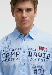 Hellblaues Hemd mit Knopfleiste und grafischen Drucken, darunter ein Bootsmotiv, eingestickter Schriftzug und ein aufgenähter Wimpel-artiger Patch auf der Brust.