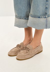 Beige suede loafers med snørebånd og metaldetaljer. Har en rund tå og flad gummisål. Sammen med lyseblå brede bukser.