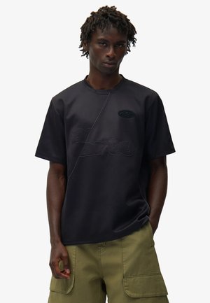 Giovane uomo con dreadlocks corti, indossa una t-shirt nera con grafica e pantaloncini cargo verde oliva, in piedi con una mano in tasca.