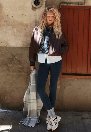 Giacca bomber marrone, leggings fitted blu navy, coperta a quadri grigia e bianca, sneakers bianche e magliette sovrapposte con dettagli del logo. Sfondo con parete texture.