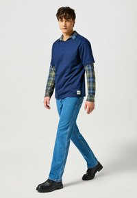 T-shirt navy sovrapposto a una camicia a quadri verde e blu, abbinato a jeans blu e scarpe nere. Stile casual a strati distintivo.