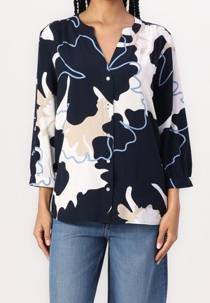 Kvinde iført en langærmet bluse med knapper, dekoreret med abstrakte blomsterprint i sort, hvid, beige og blå, kombineret med blå jeans.