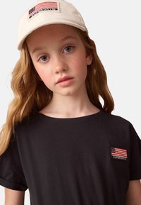 Svart bomullströja med en liten broderad amerikansk flagga och "SUPERDRY" under, matchad med en krämfärgad keps.