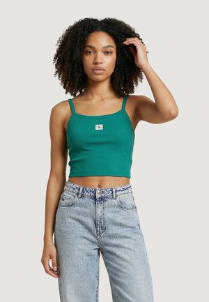 Donna con capelli ricci che indossa un top verde corto e jeans azzurri a vita alta, in piedi davanti a uno sfondo semplice.