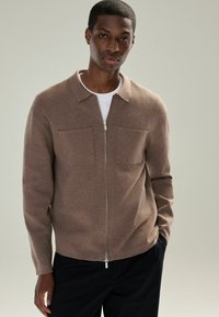Brun zip-up cardigan med krave, to brystlommer og blød tekstur, båret over en hvid t-shirt.