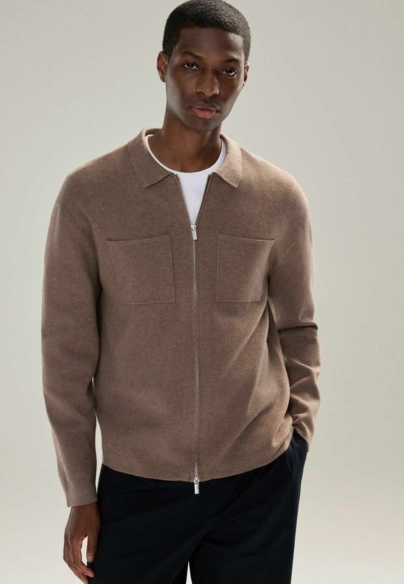 Brun zip-up cardigan med krave, to brystlommer og blød tekstur, båret over en hvid t-shirt.