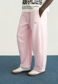 Άτομο που φοράει ροζ παντελόνι φόρμας Adidas και λευκά αθλητικά παπούτσια, στέκεται σε μπλε χαλί με το ένα χέρι στην τσέπη.