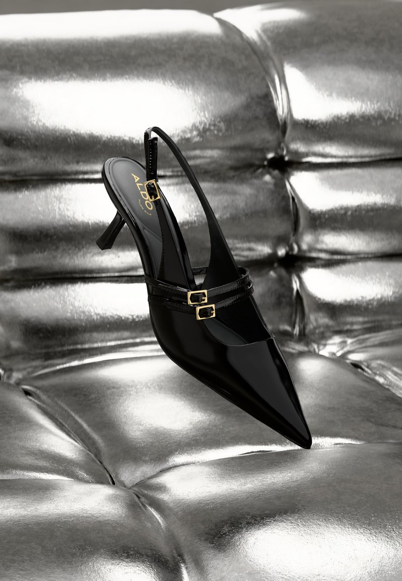 Tacco slingback in pelle verniciata nera con punta affusolata, caratterizzato da due fibbie dorate e un tacco a stiletto basso, su uno sfondo metallico argento.
