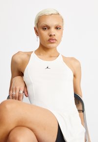 Bijela bezrukavna majica s okruglim izrezom, koja sadrži crni Jumpman logo. Glatka tekstura i prilagođeni dizajn, u kombinaciji s crnim kratkim hlačama.