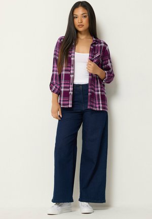 Mujer con cabello largo y oscuro está de pie vistiendo una camiseta de tirantes blanca, una camisa de cuadros morada, vaqueros oscuros de pierna ancha y zapatillas blancas sobre un fondo liso.