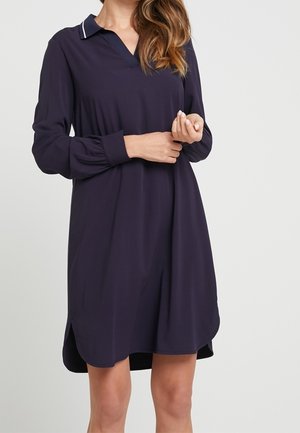 Femme portant une robe à manches longues bleu marine avec un col bordé de blanc, debout les bras légèrement croisés devant un fond uni.