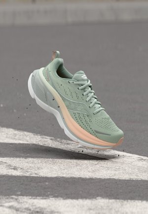 Világoszöld futócipő bézs és fehér talppal, amely az aszfalt fölött lebeg, fehér zebrák közelében.