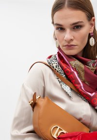 Brun läderaxelväska med en guldlogotyp, lager av färgglad silkscarf med leopardmönster och röda accenter, bärs över en beige topp.