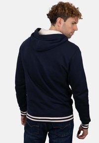 Sudadera con capucha azul marino, forro blanco y puños y dobladillo a rayas. La tela parece suave; diseño sencillo y deportivo. Vista trasera mostrada.