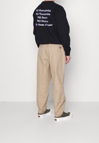 Personne portant un sweat-shirt noir avec un texte anti-discrimination au dos, un pantalon beige et des baskets vertes, debout devant un fond blanc.