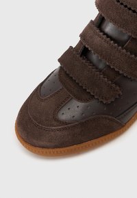 Tuvo plāns brūnu zamšādas un ādas sporta apavu purngals ar Velcro siksnām un gumijas zoli uz baltā fona.