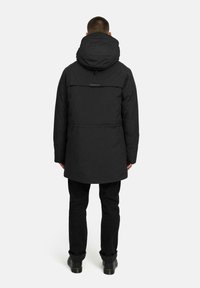 Mustapintainen huppuparka, jossa on teksturoitu pinta, keskipitkä muotoilu, takatasku ja säädettävä huppu. Yhdistetty mustiin housuihin ja saappaisiin.