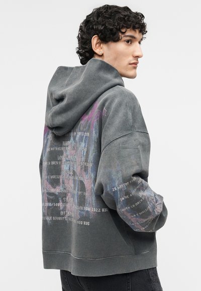 Mladá osoba s kučeravými vlasmi v nezvyčajne veľkej sivej mikine s viacfarebným grafickým textom na chrbte a rukáve, otočená bokom.