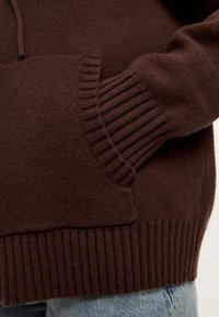 Suéter de punto marrón con puños y dobladillo acanalados, que presenta un bolsillo frontal. El material parece suave y texturizado para brindar calor.