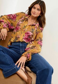 Bluse aus durchscheinendem Senfgelb mit floralem Muster und puffigen Ärmeln, kombiniert mit hochgeschnittenen blauen Jeans im weiten Schnitt und braunen Absatzschuhen.