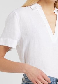 Chemise blanche en lin à manches courtes avec un col en V et coupe décontractée. Le tissu a une apparence texturée, légèrement froissée.