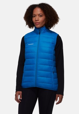 Donna che indossa un gilet imbottito blu brillante con cerniera sopra una maglia nera a maniche lunghe, in piedi davanti a uno sfondo chiaro e uniforme.