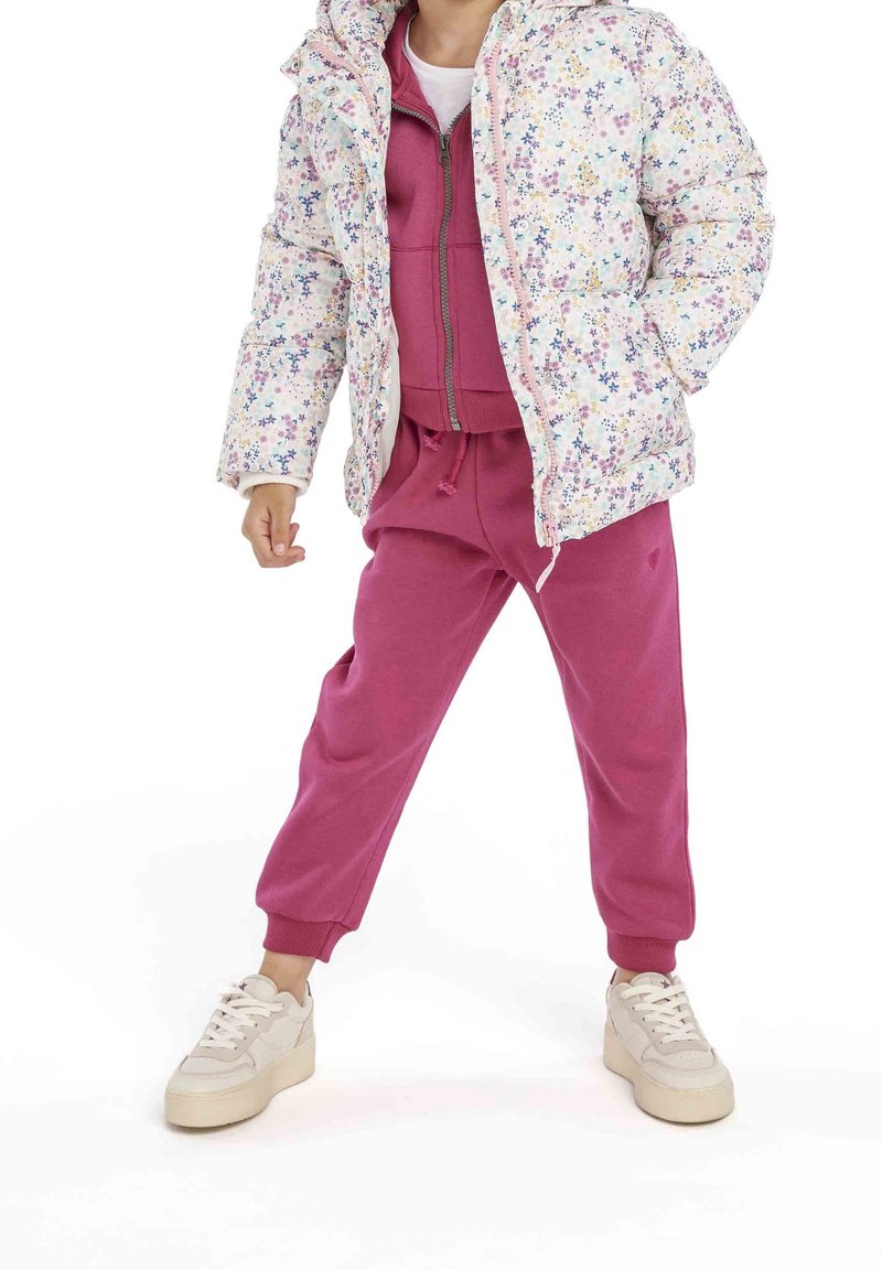 Enfant portant une doudoune blanche à motifs floraux, un sweat à capuche rose et un jogging assorti, debout jambes écartées sur un fond blanc.