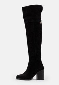 Botas de ante negras por encima de la rodilla con parte superior doblada, punta redonda y tacón grueso. Presenta un pequeño acento de logo dorado.