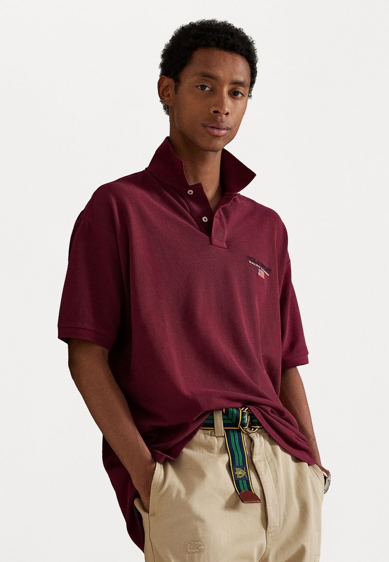 Burgundia poloshirt lühikese kraega, esipainutuste ja külgslitsidega, koos helesiniste pükste ja triibulise vööpaariga.
