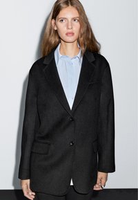 Giovane donna con capelli lunghi che indossa un blazer nero oversize su una camicia button-up azzurra, in piedi davanti a uno sfondo semplice.