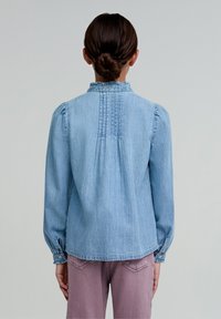 Jasnoniebieska koszula z denimu z falbaniastym kołnierzykiem, długimi rękawami i plisami z tyłu. Posiada guzikowe mankiety i miękką teksturę.