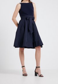 Mörkblå klänning med figurnära överdel, utsvängd kjol och en rosett i midjan. Bärs med svarta klacksandaler.