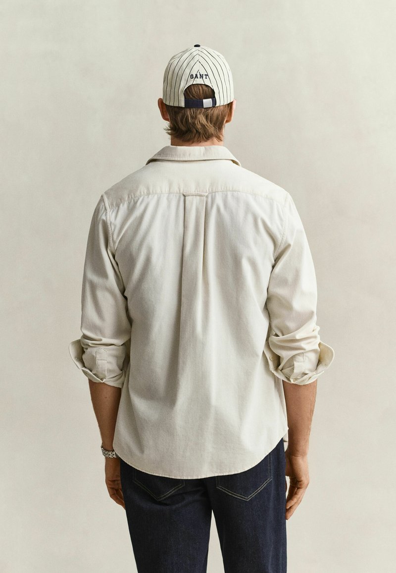 Man wearing a light beige long-sleeve shirt and striped white cap facing away against a plain background.
