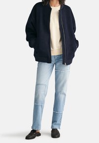 Person iført en marineblå bomberjakke over en cremefarvet sweatshirt, lyseblå patchwork jeans og sorte vævede loafers, stående med hænderne i lommerne.