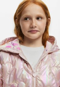 Iridescent pufferjacka med hjärtformad quiltmönster. Har ett blommigt foder, rosa dragkedja och huva. Vit skjorta under.