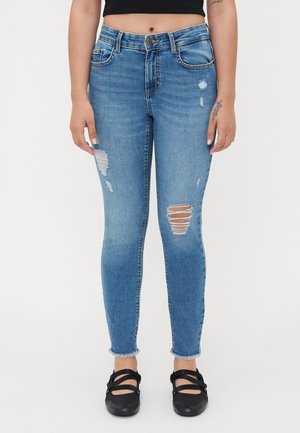 Skinny jeans in light blue denim with distressed details, featuring frayed hem and rips on the knees. Paired with black flats.