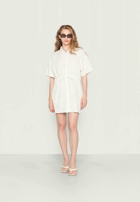 Vestido curto branco em tecido leve, com cintura amarrada, mangas curtas e um corte descontraído. Usado com óculos de sol pretos e sandálias.