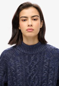 Suéter de punto azul marino con cuello alto, con patrones de trenzado texturizados y lana gruesa. Detalle deshilachado en las costuras de los hombros.