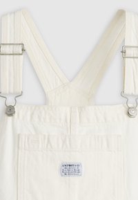 Salopettes en toile blanche avec larges bretelles, quincaillerie métallique et étiquette de marque rectangulaire sur la poche arrière. Texture lisse et conception robuste.
