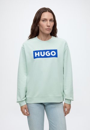 Kobieta z długimi brązowymi włosami, ubrana w jasnozieloną bluzę z niebieskim logo "HUGO" i jasnoniebieskie jeansy, stoi na białym tle.
