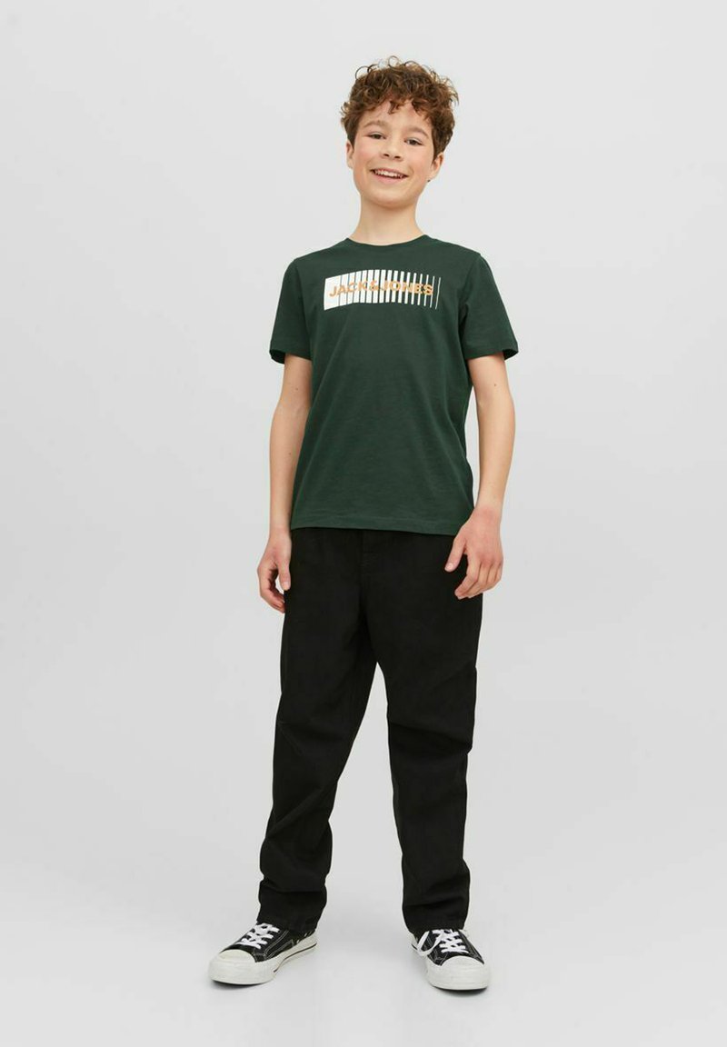 Grøn bomulds-t-shirt med et hvidt og guld grafisk design, parret med sorte bukser og sorte sneakers med hvide såler og snørebånd.
