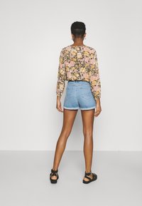 Květovaná halenka s dlouhými rukávy, růžová a žlutá, v kombinaci s lehkými džínovými šortkami. Modelka má na nohou černé sandály, vyzařující uvolněný, ležérní styl.