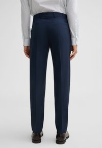 Marineblaue, taillierte Hose mit glatter Textur, Falten an der Vorderseite und zwei Gesäßtaschen. Kombiniert mit einem hellblauen gemusterten Hemd und dunklen Schuhen.