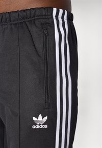 Černé sportovní kalhoty s bílým trojpruhovým designem, elastickým pasem a kapsou na zip. Měkká textura s jemným logem.