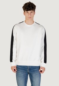 Hombre joven con cabello rizado, vistiendo un sudadera blanca con rayas negras en las mangas y jeans azules, de pie contra un fondo liso.